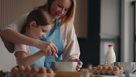 Feliz-Infancia-Y-Crianza,-La-Madre-Le-Está-Enseñando-A-Su-Pequeña-Hija-A-Cocinar-Mezclando-Ingredientes-En-Un-Tazón